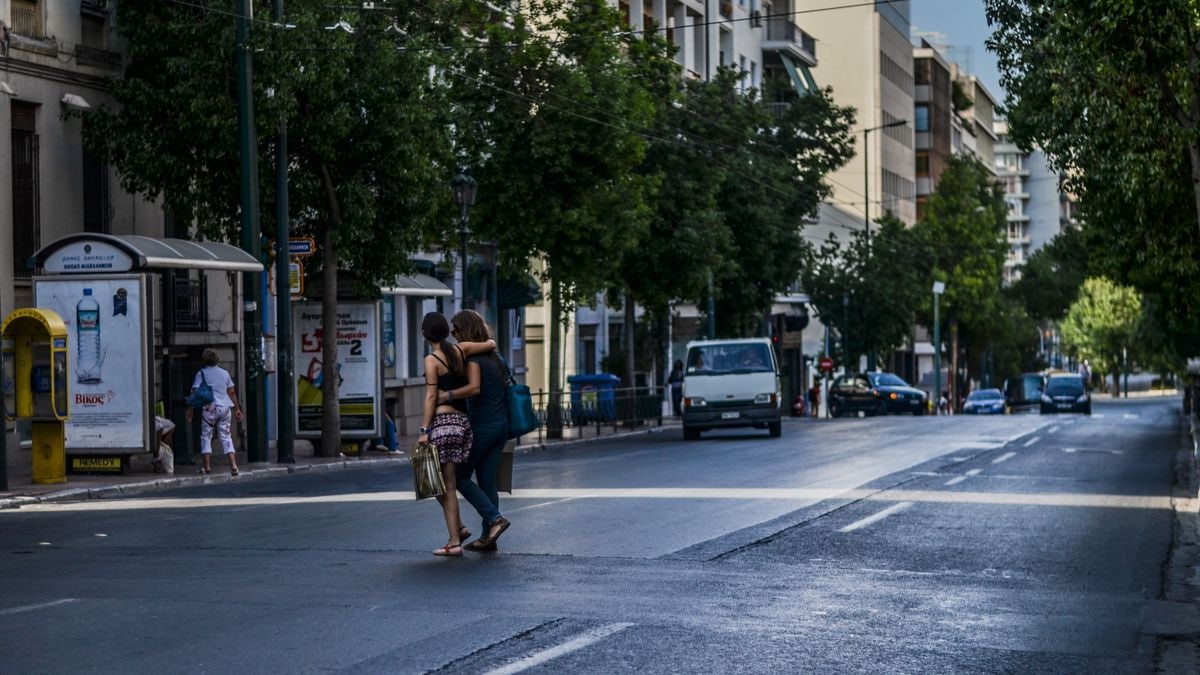 A vibrant day in Athens with people crossing a busy street lined with cars and green trees.